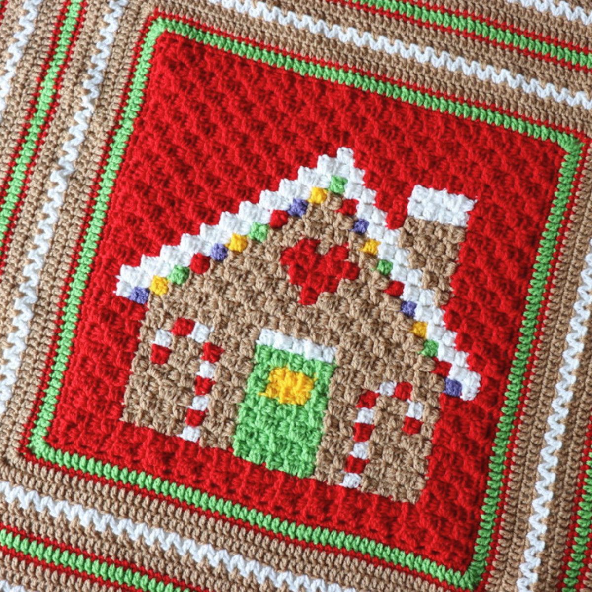 A close-up of a crocheted textile featuring a gingerbread house motif in the center, surrounded by concentric bands of red, white, green, and tan crochet.