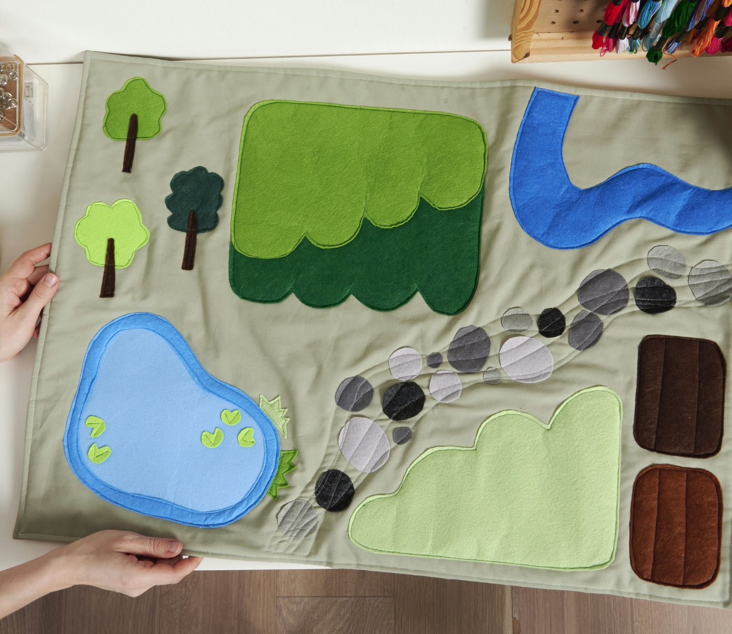 Children's play mat with green trees, blue water, and brown mountains on a wooden floor, the Coats & Clark blog post banner.