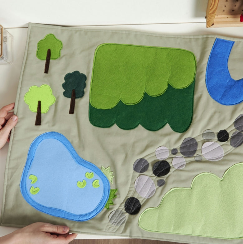 Children's play mat with green trees, blue water, and brown mountains on a wooden floor, the Coats & Clark blog post banner.