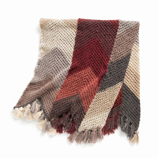 A chunky knit throw blanket with a chevron pattern in beige, rust red, brown, and dark gray, finished with fringed tassels.