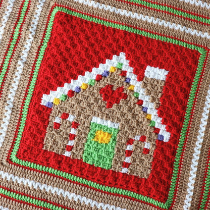 Red Heart Gingerbread House Blanket A close-up of a crocheted textile featuring a gingerbread house motif in the center, surrounded by concentric bands of red, white, green, and tan crochet.