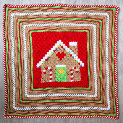Red Heart Gingerbread House Blanket A square crocheted textile featuring a central gingerbread house motif surrounded by concentric decorative borders in red, brown, white, and green on a light gray background.