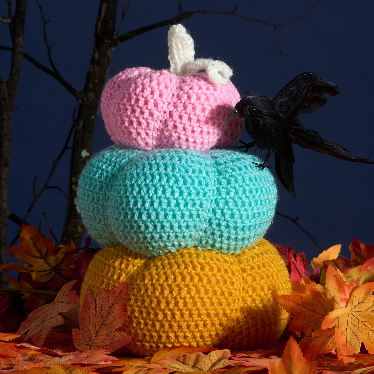 Crocheted stack of pumpkins with a crow on a dark blue background with autumn leaves.