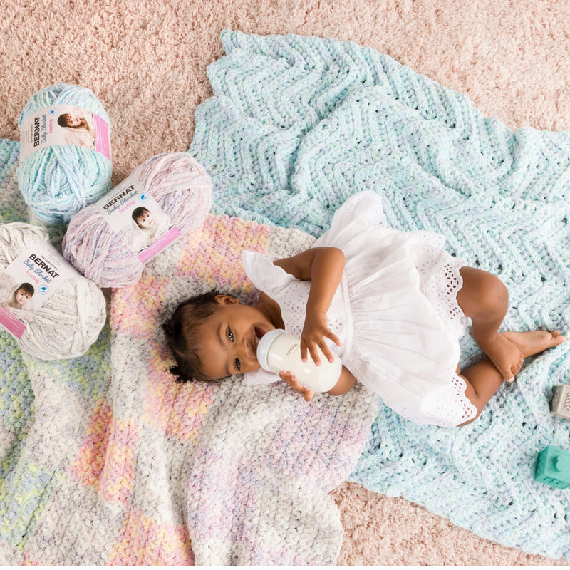 Baby in a white dress lying on a colorful crochet blankets with yarn balls in the background.