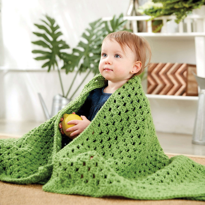 A cute baby is holding a lemon and covered with our Caron easy peasy green baby blanket made from the Caron one pound, Grass Green shade yarn.